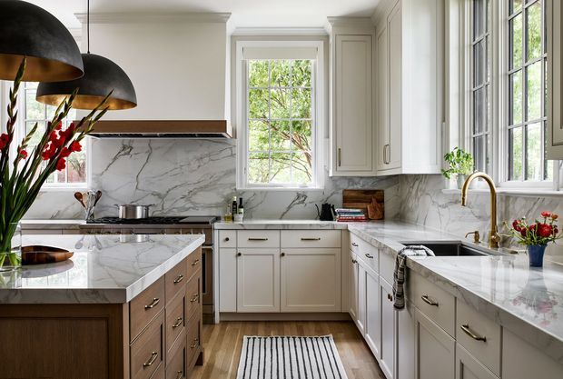 A bright, refined traditional kitchen with cream and white raised-panel cabinetry, extensive veined marble-look surfaces, a central island, large multi-pane windows, and dark dome pendant lights. The room includes a gas cooktop, sink beneath the window, brass-toned tapware and hardware, timber flooring, and a compact striped runner.