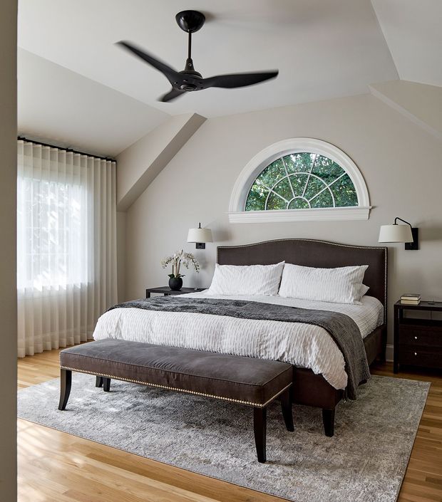 A refined bedroom with a large upholstered bed dressed in white bedding and a charcoal throw, paired with a matching nailhead-trimmed bench. The room features light timber flooring, a large grey area rug, dark bedside furniture, wall-mounted lamps, sheer floor-to-ceiling curtains, and a distinctive arched fanlight window with decorative radial muntins. A black ceiling fan and angled ceiling detail add architectural interest. The palette is neutral, calm, and softly lit.