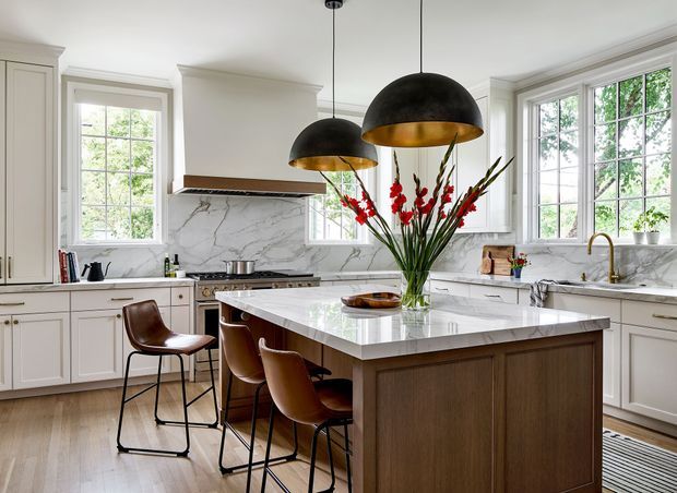Bright, spacious traditional-style kitchen featuring white shaker cabinetry, marble-look stone surfaces, a large central island with a dark timber base, integrated seating, and black dome pendant lights with brass interiors. Large divided-pane windows provide abundant natural light, while a sculptural floral arrangement forms the island centerpiece.