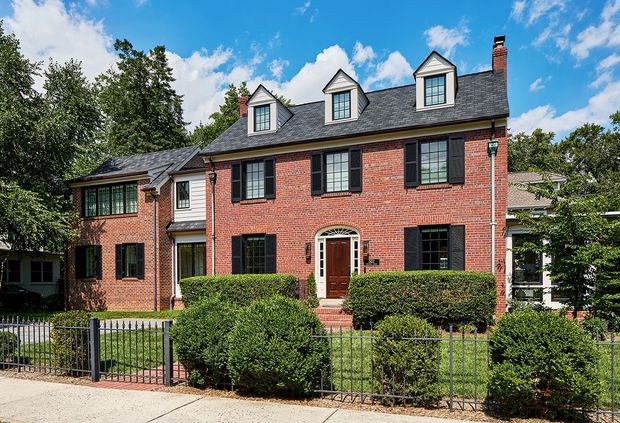 Exterior view of a substantial traditional red-brick multi-storey residence with a steep dark shingle roof, three front dormer windows, black shutters, white-trimmed windows, a central arched timber entry door, and a landscaped front garden behind a metal fence. The house has a connected side wing and sits in a leafy suburban setting under a bright blue sky.