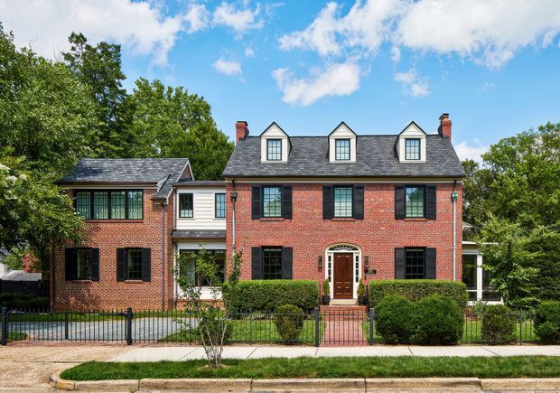 Front exterior view of a traditional red-brick multi-storey residence with a symmetrical main facade, dark shutters, a slate-like gabled dormer roof, brick chimneys, and a central arched entry door. The house sits behind a low metal fence with clipped hedges and a paved pedestrian path, surrounded by mature trees under a bright sky.