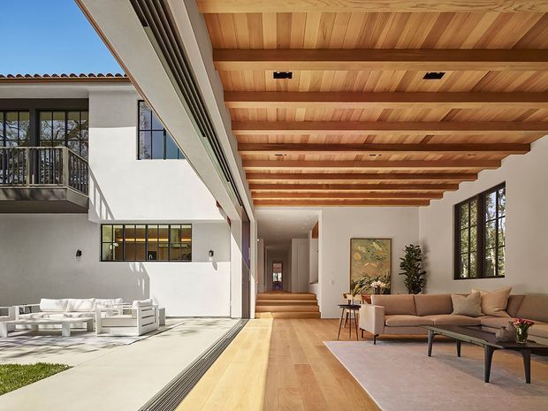 Sunlit contemporary living room opening onto an internal courtyard, with a warm exposed timber beam ceiling, pale timber flooring, white walls, expansive black-framed glazing, and a neutral sofa and coffee table. A stair and hallway recede through the centre, while an adjacent upper-level balcony and courtyard are visible on the left.
