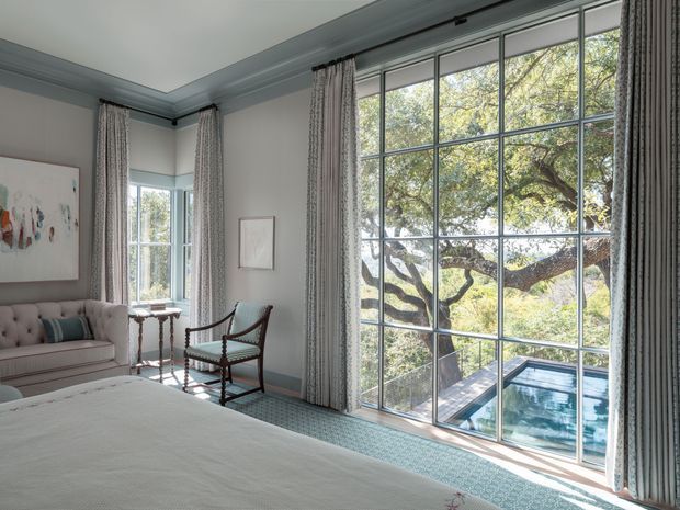 Elegant bedroom with a large upholstered bed, tufted seating, a wood-framed armchair, patterned floor-to-ceiling curtains, and expansive black-framed gridded windows overlooking a leafy garden and swimming pool. The room uses a restrained pale blue-grey and cream palette with traditional detailing and abundant natural light.