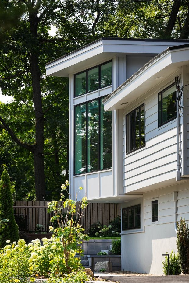 Exterior view of a contemporary multi-level house set among mature trees and landscaped garden beds. The building has crisp white horizontal cladding, dark-framed expansive glazing, projecting flat roof planes, and a partially visible stepped garden and paved approach.