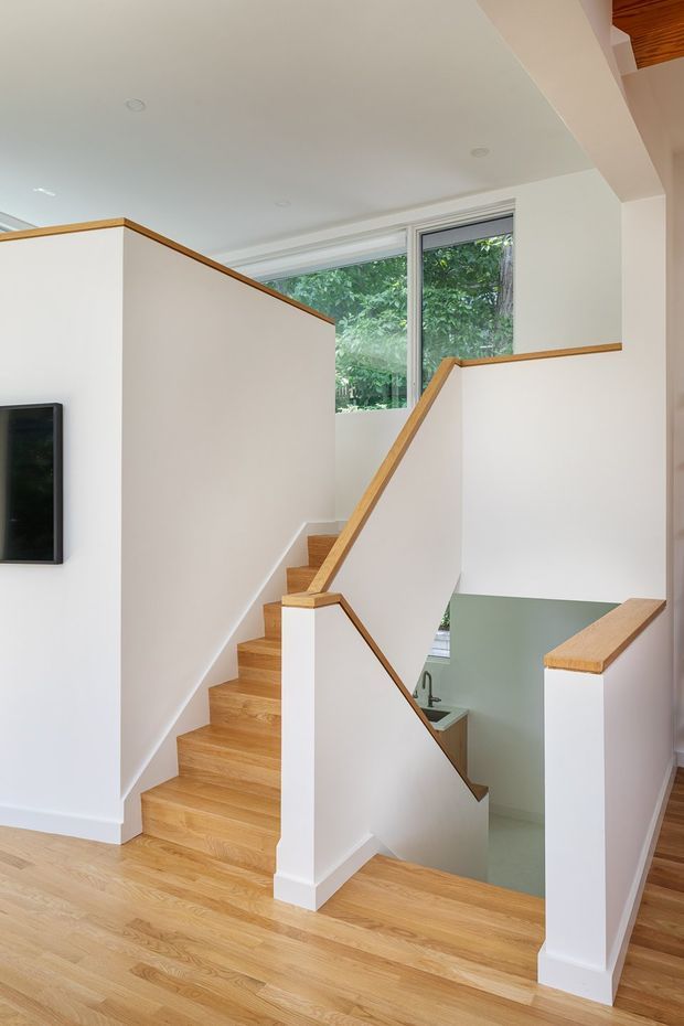 Bright contemporary interior view of a timber staircase and stair landing. The staircase has white painted walls and balustrade walls with warm light-wood handrails and treads, opening toward a lower level. Large windows bring in greenery and daylight, while a small portion of timber ceiling detail is visible at the upper right.
