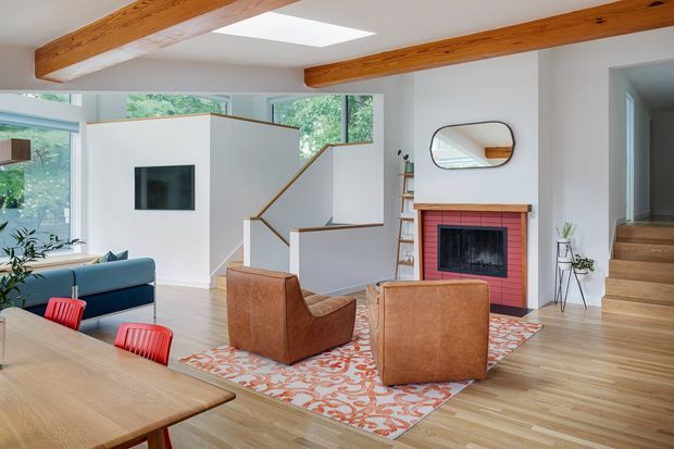 A bright, open-plan living and dining area with exposed timber ceiling beams, large windows, and a skylight. The room features a blue sofa, leather armchairs, a timber dining table with red chairs, a patterned rug, and a red-painted brick fireplace. A sculptural white stair enclosure leads to a timber staircase, with a hallway visible to the right.