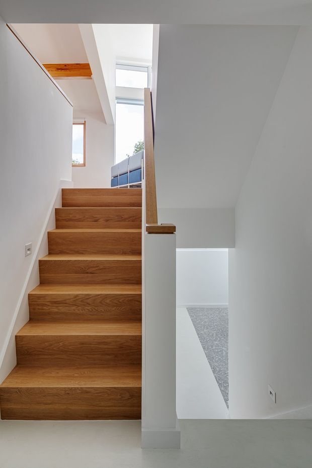 A bright, minimalist interior stairway with broad solid timber stair treads and risers, white painted walls and structural partitions, and a simple timber handrail. The stair rises toward a landing with tall clerestory-style glazing and a glimpse of an upper-level balcony or railing. A lower stair or void area is visible to the right, with a pale patterned floor surface. The composition is clean and architectural, emphasizing the stair geometry, natural wood grain, and abundant daylight.