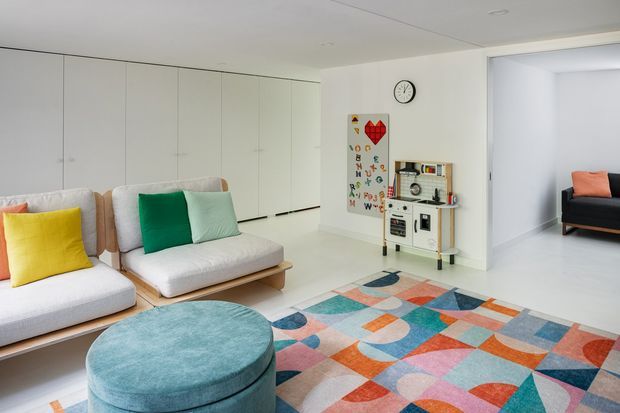 Bright, open-plan living room with a minimalist white built-in storage wall, pale upholstered seating, a large rounded teal ottoman, and a colourful geometric area rug. A children's play and storage unit sits against the wall, while an adjacent room is visible through a wide opening.