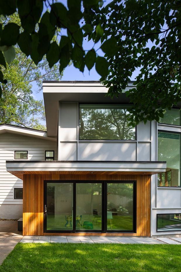 Exterior view of a contemporary multi-level residence set among mature trees. The facade combines pale horizontal cladding and smooth light wall panels with expansive dark-framed glazing. A projecting upper volume shelters a ground-level glazed opening framed in warm vertical timber cladding, creating a clear contrast between the white facade and natural wood. A lawn and paved edge are visible in the foreground.