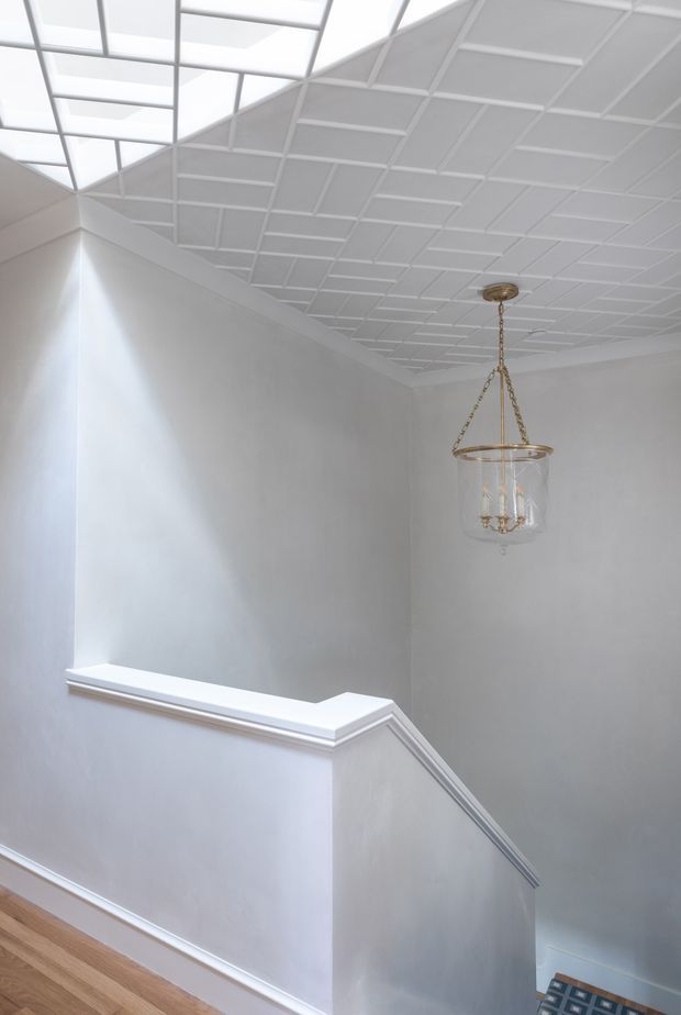 Bright, refined stairway or landing with pale grey walls, white decorative trim, a white coffered or geometric ceiling, and a large skylight providing abundant natural light. A traditional glass-and-brass pendant hangs above the stairwell, while light timber flooring and patterned stair carpeting add warmth.