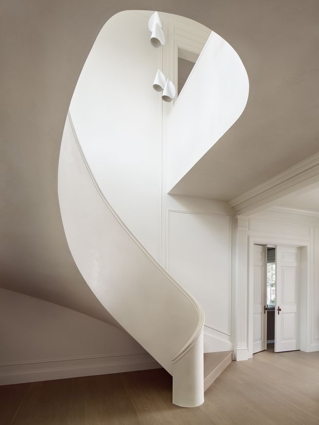 A refined interior stairway featuring a sculptural curved white staircase with a smooth solid balustrade and rounded newel, set against white paneled walls and ornate classical door and ceiling trim. Pale timber flooring and soft neutral tones create a bright, elegant entry circulation space.