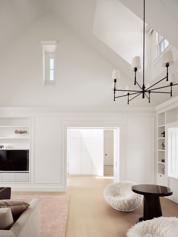 Bright, refined living room with white panelled walls, built-in shelving and media cabinetry, pale timber flooring, and a high vaulted ceiling. A striking black multi-arm chandelier hangs above a round dark side table, while curved white bouclé lounge chairs and a neutral sofa sit on a pale rug. A wide opening leads to a light-filled adjoining space, and tall windows bring in abundant natural light.