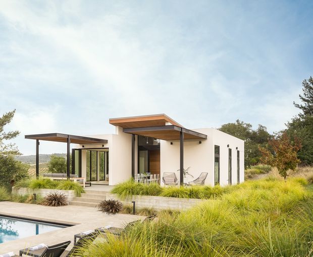 Exterior view of a low-profile contemporary house set within landscaped grasses and trees. The building has white rendered walls, extensive floor-to-ceiling glazing, projecting flat roof planes with exposed timber-toned soffits, and a covered outdoor sitting area. A swimming pool and paved terrace are visible in the foreground, with a gently sloping natural landscape beyond.