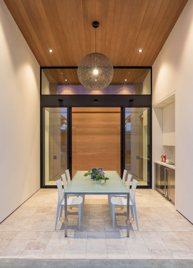 A refined dining room with a long rectangular glass dining table and white chairs, positioned beneath a large spherical pendant light. The room features a warm timber-lined vaulted ceiling, pale tiled flooring, crisp light walls, and black-framed glazed doors opening to a timber-lined space beyond. A slim drinks cabinet is integrated into the right wall, while concealed and recessed lighting adds a calm, contemporary ambience.