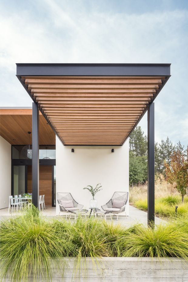 A contemporary outdoor terrace beneath a large timber-slatted pergola supported by dark metal posts. The covered patio contains two lounge chairs and a small table, with a dining setting visible to the left through the open connection to the house. White rendered walls, warm timber soffit and wall cladding, dark-framed glazing, and layered ornamental grasses define the landscaped residential exterior.