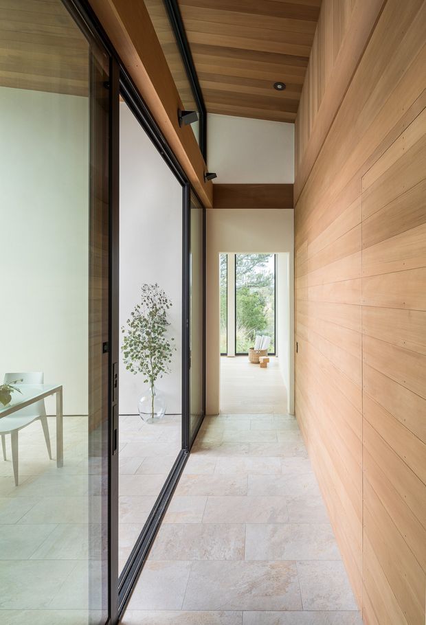 A narrow, refined interior hallway with pale stone tile flooring, extensive warm timber wall and ceiling lining, exposed timber beams, and black-framed full-height glazing on the left. The glazing reflects a minimalist sitting or dining area, while the corridor leads toward a bright room with a large window and outdoor greenery beyond. Recessed and track lighting provide understated illumination.