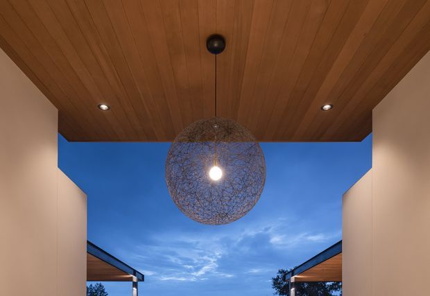 Architectural view from beneath a covered outdoor opening, featuring a warm timber-lined ceiling with recessed downlights and a large spherical woven pendant light. Pale side walls frame an open view of the blue evening sky, with portions of adjacent rooflines visible on either side. The composition emphasizes a sheltered outdoor threshold or courtyard-like space and a strong indoor-outdoor connection.