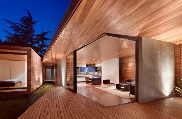 Dusk view of a contemporary timber-clad residence opening onto a broad timber deck and courtyard. The image is dominated by a warm timber soffit and decking, large dark-framed glazed openings, and a raw concrete feature wall. Through the open glazing, a minimalist living area with low seating and a kitchen or dining zone is visible. A separate bedroom-like volume is visible at left, connected by the landscaped courtyard and deck.