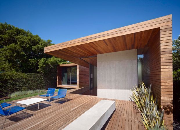 Exterior view of a contemporary single-storey home opening onto a spacious timber deck. The building features a strong cantilevered flat roof, extensive warm timber cladding and soffits, a smooth concrete feature wall, and large glazed openings. Blue outdoor seating and built-in concrete-and-timber deck elements create a relaxed outdoor living area surrounded by established vegetation.