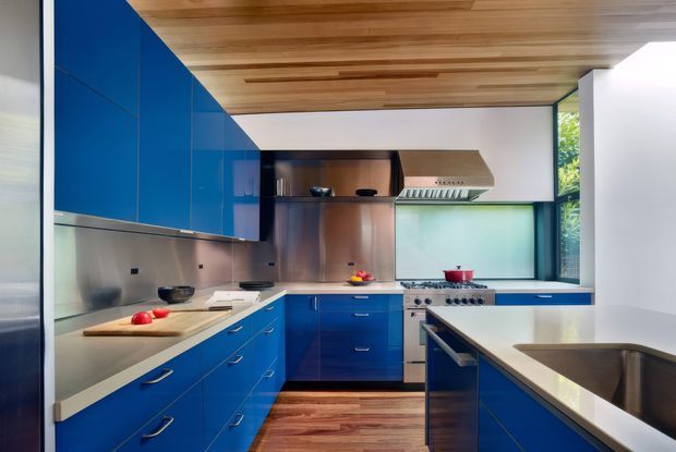A contemporary galley-style kitchen with vivid blue full-height cabinetry, stainless-steel appliances and splashback, pale stone benchtops, and a warm timber-lined ceiling. A large island with an integrated sink occupies the foreground, while a stainless rangehood, cooker, and tall window complete the compact but well-equipped space.