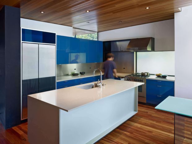 A contemporary kitchen with a large central island, glossy deep-blue cabinetry, integrated appliances, stainless-steel cooking and ventilation equipment, and a pale stone-look island benchtop. Warm timber ceiling lining and timber flooring contrast with the cool blue joinery. Recessed downlights, extensive glazing, and a partially visible dining table complete the space.