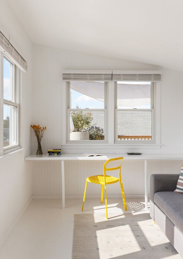 Bright, compact white-lined room with a built-in desk beneath multiple windows, a vivid yellow chair, a grey upholstered sofa edge, and a pale patterned rug. White vertical wall panelling, white-painted trim, roller blinds, and strong natural sunlight create an airy workspace or reading nook.