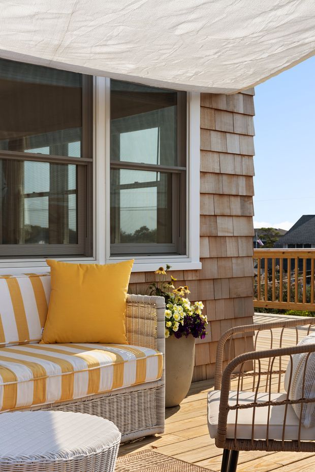 A bright coastal-style outdoor terrace or balcony with a woven wicker seating arrangement, yellow-and-white striped cushions, a round wicker ottoman, potted flowers, timber decking, and a timber balustrade. The adjacent building has shingle cladding, large framed windows, and a light textured overhead canopy or ceiling. The scene is sunlit, relaxed, and distinctly seaside-inspired.