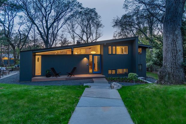 A compact modern dark-clad residence set among mature trees, photographed at dusk. The single-storey building has a low-pitched roof, large warm-lit windows, a recessed covered porch with outdoor seating, and a concrete path leading through a lawn to the entrance.