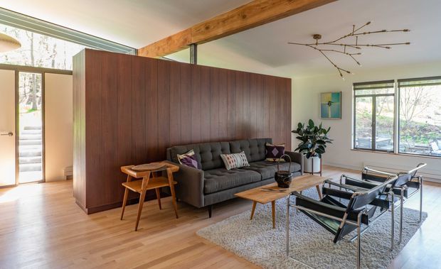 A bright mid-century-inspired living room with a dark timber-paneled partition wall, exposed timber ceiling beam, large glazed openings, a charcoal sofa, leather sling chairs, and low timber coffee tables arranged over a textured rug. The room opens toward a glazed exterior doorway and landscaped surroundings.