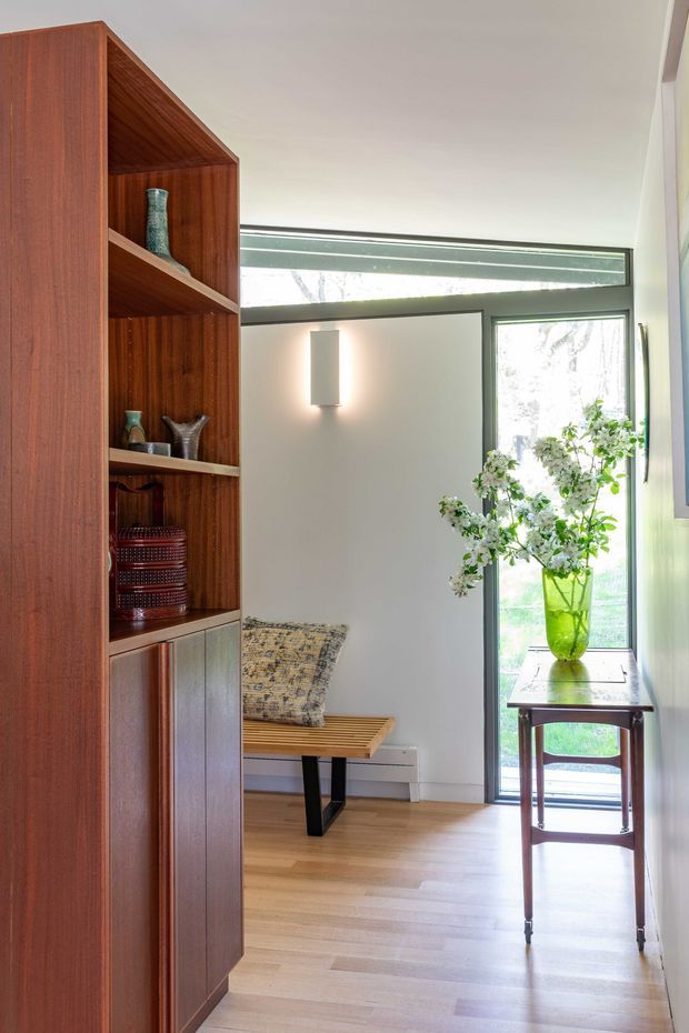 A bright, refined interior vignette featuring a tall built-in dark timber shelving and cabinet unit with open display niches. A simple wall sconce illuminates the pale wall beside a glazed door and clerestory window. The room has light timber flooring, a timber bench with a patterned cushion, and a small dark timber console table holding a green glass vase with flowering branches.