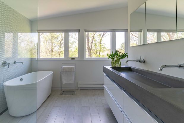 Bright contemporary bathroom with a freestanding white bathtub, long double vanity with a dark grey benchtop, wall-mounted chrome tapware, pale tiled flooring, and a small-format textured tile feature wall. Large windows and mirrored cabinetry create an airy, light-filled space.