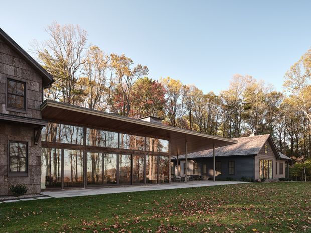 Exterior view of a contemporary multi-wing residence set among mature deciduous woodland. The composition features a dark textured masonry-clad building at left, a long flat-roofed glazed connector with extensive floor-to-ceiling windows, and a lower pitched-roof wing with dark horizontal cladding at right. A broad lawn and autumn trees surround the house.