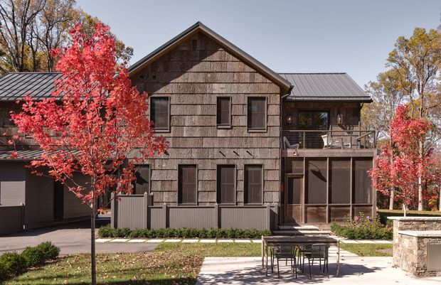 Exterior view of a multi-storey residential building with a prominent front-facing gable, dark grey metal pitched roofs, weathered timber shingle cladding, and dark-framed windows. A covered balcony and screened lower-level outdoor area sit on the right side, with a paved patio, dining table, low stone outdoor structure, lawn, and autumn trees in front.