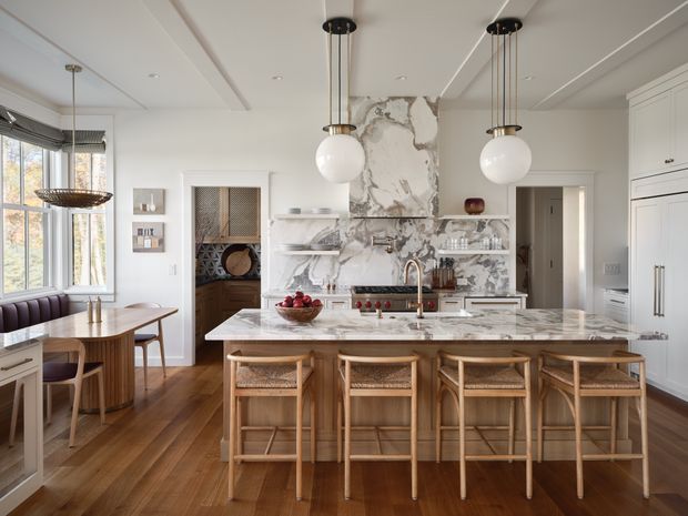 A refined, spacious kitchen with a large marble island and four woven-seat timber bar stools. The room features white cabinetry, a dramatic veined marble splashback and benchtop, warm timber flooring, globe pendant lights, integrated appliances, and an adjoining dining nook with a curved banquette by the windows. The overall scheme is bright, warm, and classic-contemporary.