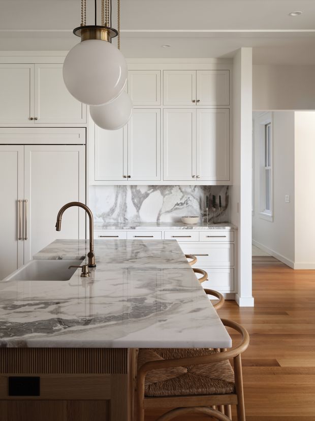 A refined, bright kitchen viewed from the island. White framed cabinetry surrounds a large veined stone island and matching backsplash, with an integrated sink and curved bronze-toned faucet. Woven-seat timber bar stools line the island, while globe pendant lights hang above. Warm timber flooring continues into the adjacent hallway, creating a calm, high-end interior.