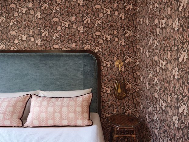 A close-up view of a richly patterned bedroom corner featuring a bed with a blue upholstered headboard, white bedding, and blush-pink patterned cushions. The walls are covered in dense botanical wallpaper in muted brown and taupe tones. A small dark wood bedside table and a brass-toned wall light add vintage character.
