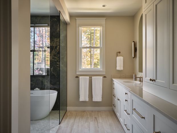 A bright, refined bathroom viewed along a long layout, featuring a freestanding white bathtub behind a frameless glass shower enclosure, a double vanity with extensive paneled built-in cabinetry, a pale stone countertop, white trim, and warm light-toned wood-look flooring. A central window brings in natural light and views of trees, while brushed metal hardware and white towels provide understated detailing.