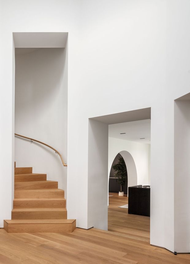 A minimalist interior hallway and stairway with white plaster walls, warm natural timber flooring, and a straight staircase leading upward along the left wall. The staircase has timber treads and a simple curved timber handrail. A large rectangular doorway opens into an adjoining room with matching timber flooring, dark built-in cabinetry or furniture, an indoor plant, and a rounded arched opening. The composition is clean, light, and architecturally focused, with recessed ceiling lighting and strong geometric openings.