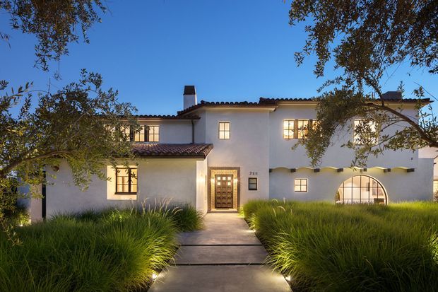 Exterior dusk view of a substantial white stucco residence with a red clay tiled roof, projecting wings, dark-framed multi-pane windows, a timber entry door, and a distinctive arched ground-floor window. A straight paved walkway passes through landscaped ornamental grasses and mature trees toward the entrance. The home has a warm Mediterranean or Spanish-influenced character.