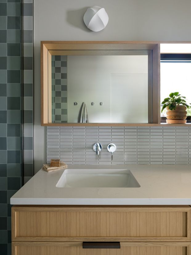 A contemporary bathroom vanity viewed straight on, featuring a wall-mounted timber-framed mirror, pale integrated basin, light stone benchtop, fluted timber cabinetry, grey-green tiled walls, chrome tapware, and a small potted plant on the sill. A dark green tiled shower wall is visible at the left edge. The clean geometric composition has warm timber balanced with cool muted tiles.
