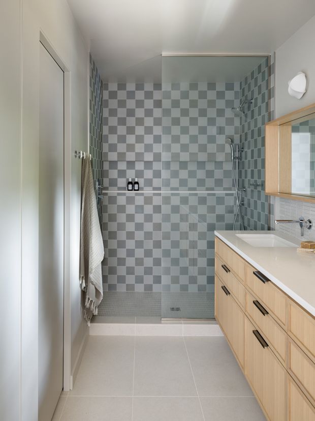 A compact contemporary bathroom viewed from the entrance, featuring a walk-in shower enclosed by clear glass, grey-and-white checkerboard wall tiles, matching small-format shower floor tiles, large light-grey floor tiles, and a pale timber floating vanity with a white integrated basin and black hardware. A mirrored cabinet and wall light are visible on the right.