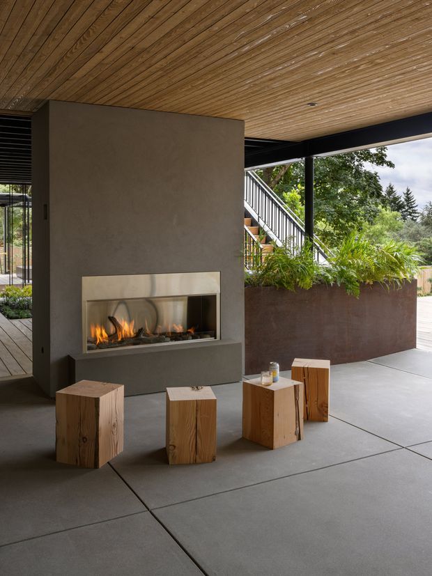 A covered, open-sided outdoor living area with a large built-in fireplace, broad concrete floor slabs, a warm timber slatted ceiling, and black structural framing. Rustic solid timber cube stools form a casual seating arrangement around the fireplace, while a raised rust-toned planter provides greenery beside a deck and stair connection.