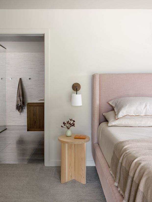 A softly styled bedroom with a pale pink upholstered bed and layered neutral bedding. A curved light-wood bedside table, wall-mounted white sconce with timber backing, and small ceramic vase create a calm, refined setting. An open doorway reveals an adjacent tiled bathroom with a timber vanity and wall-mounted towel.