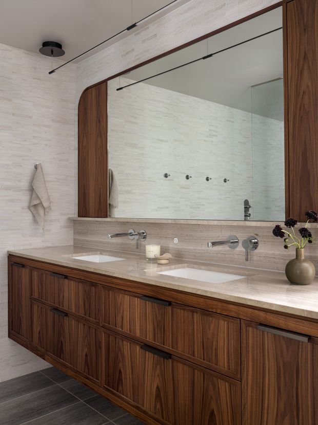 A refined double vanity bathroom with a long floating-style dark timber vanity, two integrated white basins, pale stone benchtop, wall-mounted chrome tapware, and a large mirrored cabinet. The walls are clad in pale textured stone or tile, with slim black linear lighting and warm timber detailing creating a contemporary, luxurious feel.