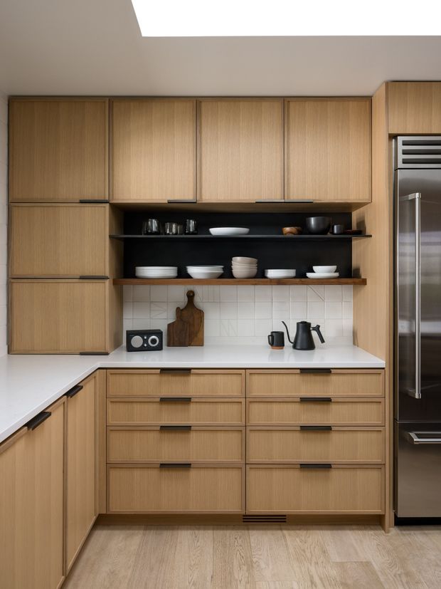 A compact, carefully detailed kitchen with full-height light timber cabinetry, integrated drawer fronts, open black display shelving, a white countertop, and a white tiled backsplash. A stainless-steel refrigerator is visible on the right, with black small appliances and neutral crockery arranged on the shelves and bench. The space is illuminated by a large ceiling opening or skylight and has a warm, refined contemporary appearance.