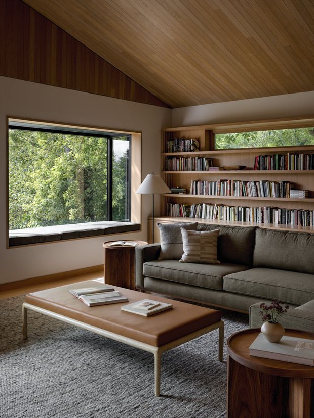 A warm, mid-century-inspired living room with a large picture window overlooking greenery, extensive built-in timber bookshelves, a grey upholstered sofa, and a low leather-topped coffee table. The room features a dramatic sloped timber-lined ceiling, timber wall panelling, neutral textiles, and dark wood occasional tables, creating a refined and cozy reading and sitting space.
