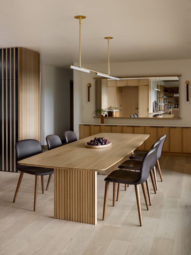 A warm, refined dining area centered on a large rectangular timber dining table with a fluted pedestal base and upholstered dark dining chairs. Pale timber flooring, integrated timber cabinetry, vertical timber screening, a mirrored open-shelving niche, and slim brass-and-white pendant lights create a cohesive contemporary interior. A kitchen or serving joinery zone is visible behind the dining area.