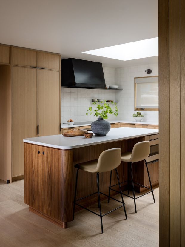 Warm, refined contemporary kitchen with a substantial island clad in rich vertical-grain timber and topped with a white benchtop. Integrated timber cabinetry lines the left wall, while a black sculptural rangehood contrasts with the white square-tile splashback. Two upholstered counter stools sit at the island, and a large skylight brings in natural light. The room has a calm, earthy palette with carefully styled greenery and ceramics.