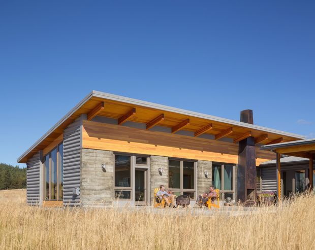 Single-storey contemporary timber-and-concrete house set in a dry grassy landscape. The building has a broad low-pitched roof with exposed timber rafters and deep eaves, warm timber soffits and beams, horizontal dark cladding, large glazed openings, and a covered outdoor terrace with a prominent dark structural post and chimney.