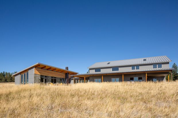Wide exterior photograph of a contemporary multi-wing residence set in a dry grassy landscape. The home has horizontal timber-toned cladding, a low-pitched standing-seam metal roof, broad eaves, timber posts and beams, and extensive dark-framed glazing. A smaller flat-roofed or shallow-pitched wing connects to the larger two-level volume. Clear blue sky and surrounding trees form the background.